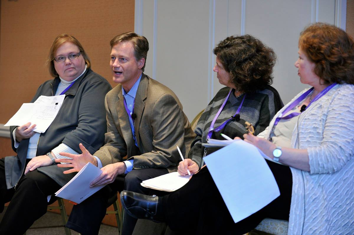 Tony, seated on a chair, facilitating a conversation. One woman on his right and two women on his left, also seated, are listening. All four people are holding pages of notes.
